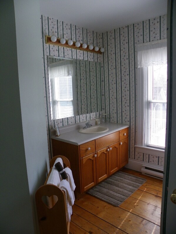 Une salle de bain avec un meuble-lavabo en bois, un évier et un miroir. On aperçoit une fenêtre avec des stores et une porte.