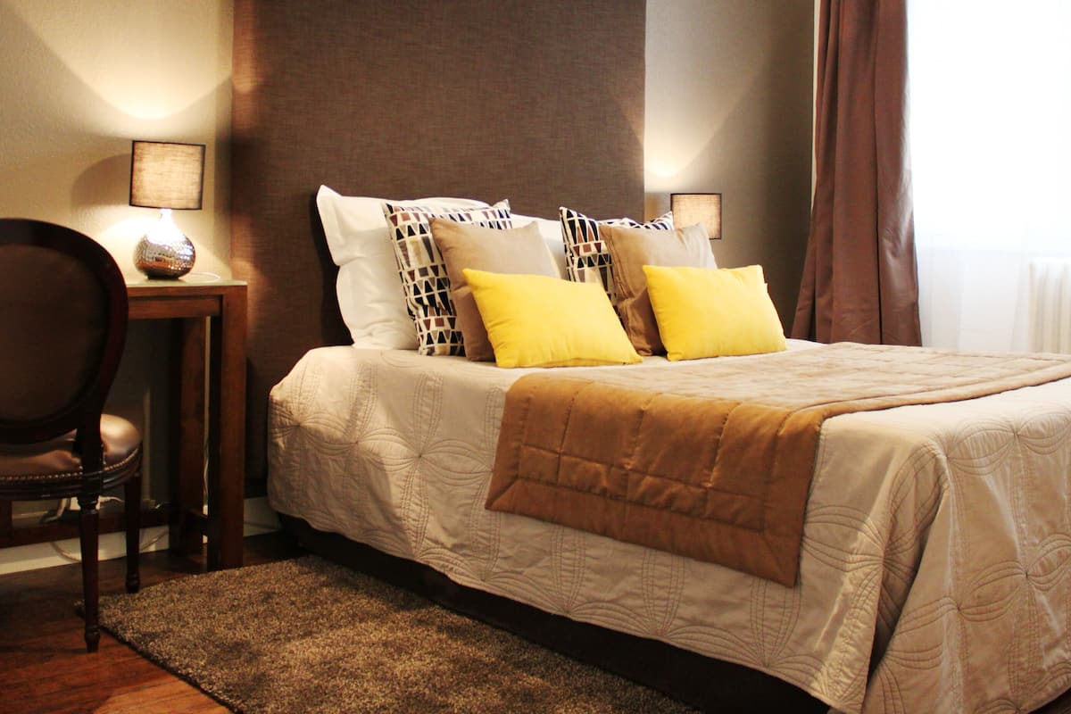 A neatly made bed with a brown blanket, a chair, a small table with a lamp, and two wall-mounted picture frames.