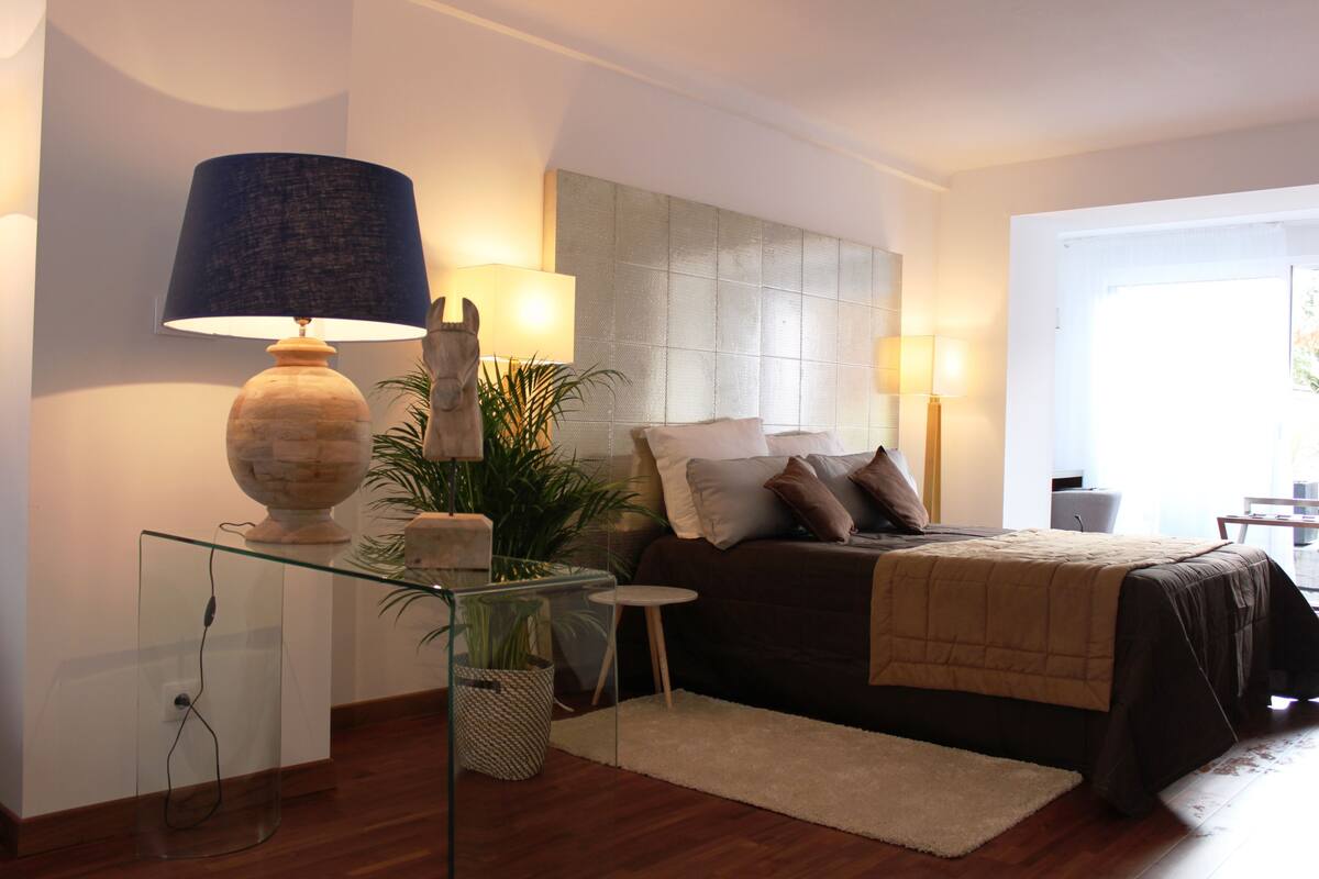 A modern bedroom with a bed, bedside lamps, a glass table with a lamp, and a potted plant.