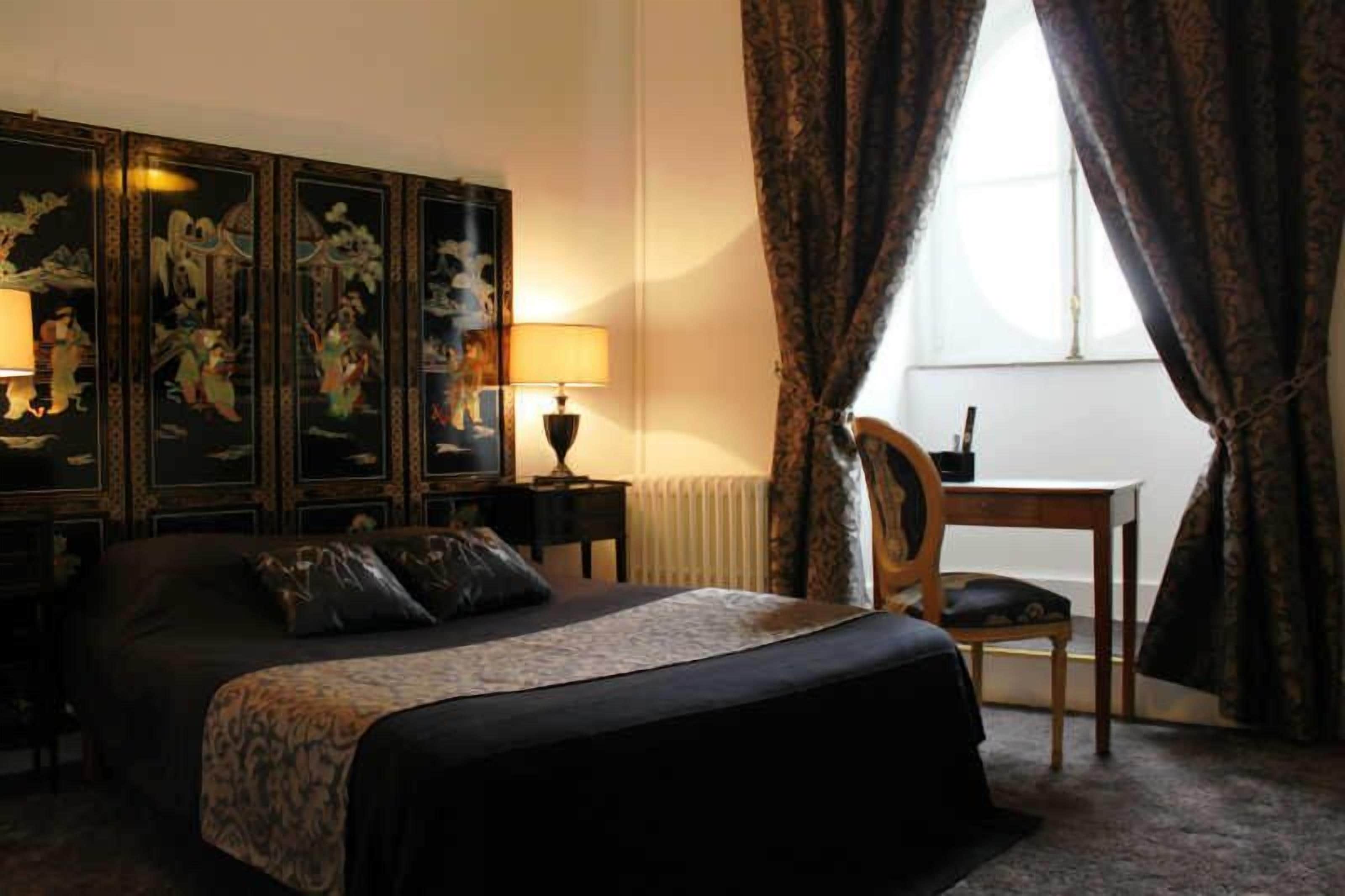 A hotel room with a large bed, two bedside tables with lamps, a wooden chair, a small table, and a large window with curtains.