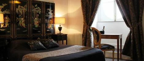 A hotel room with a large bed, two bedside tables with lamps, a wooden chair, a small table, and a large window with curtains.
