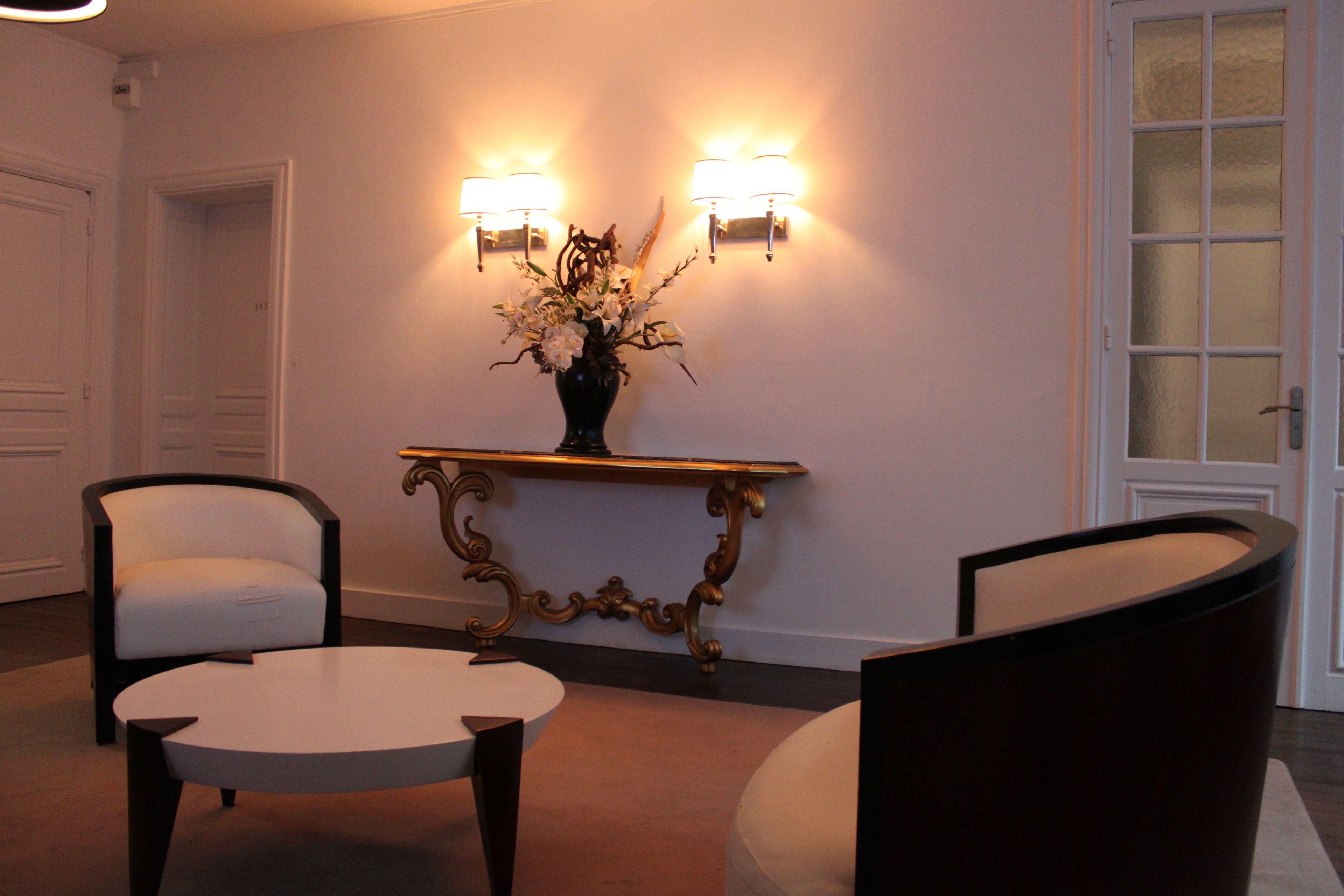 A lobby with two white chairs, a round table, a decorative vase with flowers, and wall-mounted lamps.