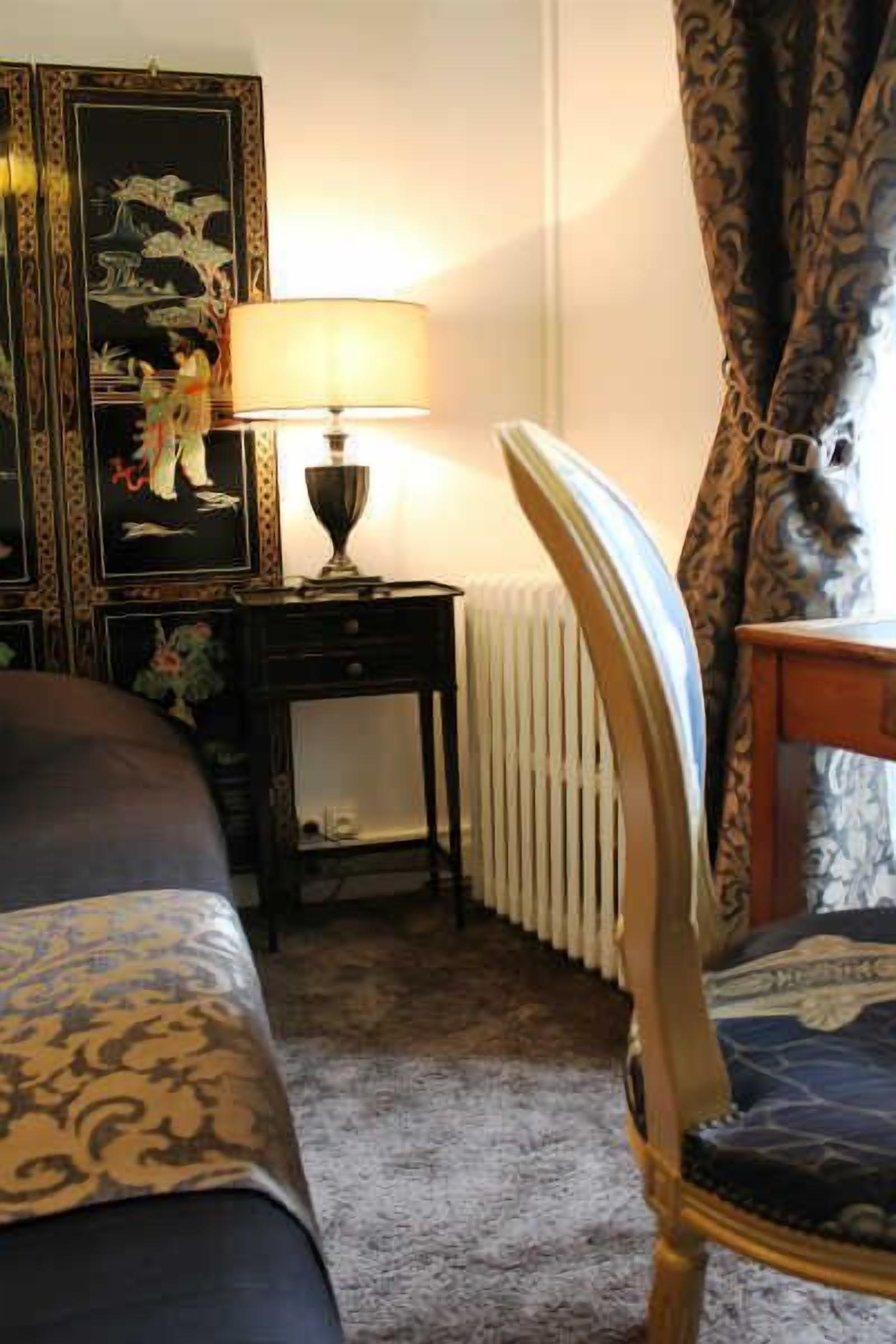 A hotel room with a bed, a table with a lamp, a radiator, and a chair.