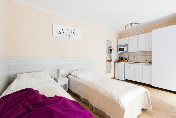 A bedroom with two beds, a kitchen area with white cabinets and appliances, and a wall-mounted artwork.