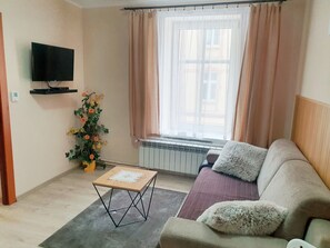 A living room with a sofa, a small table, a flat-screen TV mounted on the wall, a radiator, and a window with curtains. - Aparthouse Apartamenty (Katowice)