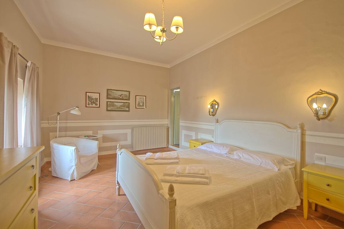 Quarto com uma cama de casal, mesa de cabeceira, cadeira, escrivaninha e abajur.