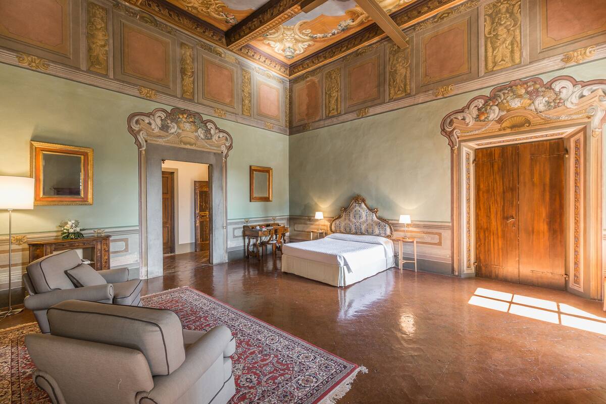 Um quarto espaçoso com uma cama grande, área de estar com cadeiras, uma mesinha e lareira.