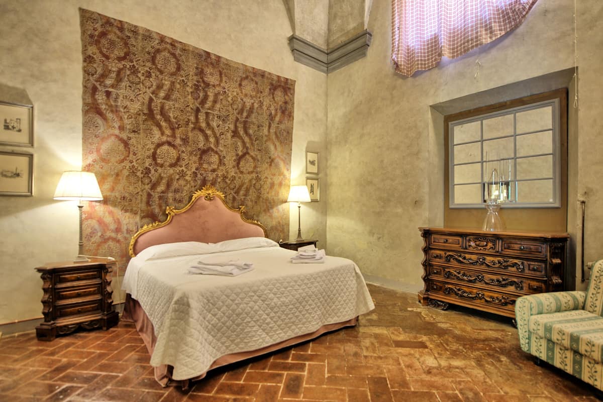 Quarto com cama grande, duas mesas de cabeceira com abajures, cômoda de madeira e tapeçaria estampada na parede.