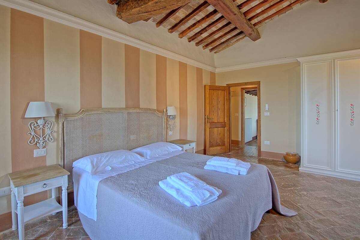 Quarto com uma cama grande, cabeceira de madeira, duas mesas de cabeceira com abajures, guarda-roupa e uma porta que dá para outro cômodo.