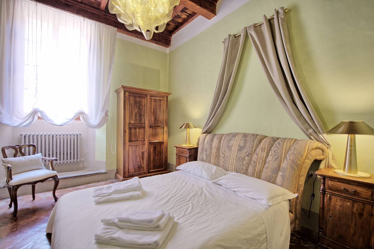 Quarto com uma cama grande, um guarda-roupa de madeira, uma cadeira, uma mesa de cabeceira com abajur e uma janela com cortinas.