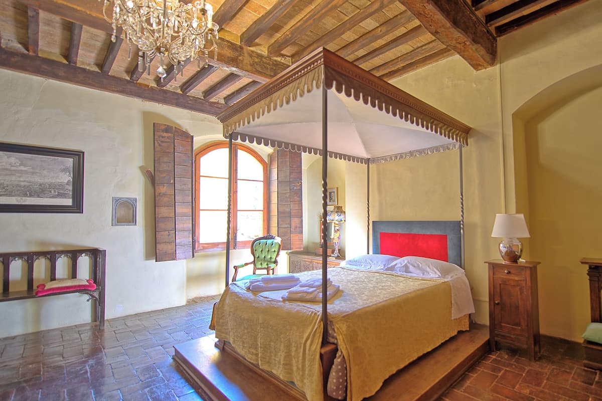 Uma cama de dossel com colunas e cabeceira de madeira, um banquinho com almofada, uma mesa pequena com abajur e um candelabro.