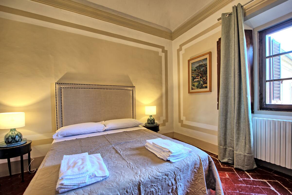 Quarto de hotel com uma cama grande, duas luminárias de cabeceira, um quadro na parede e uma janela com cortinas.