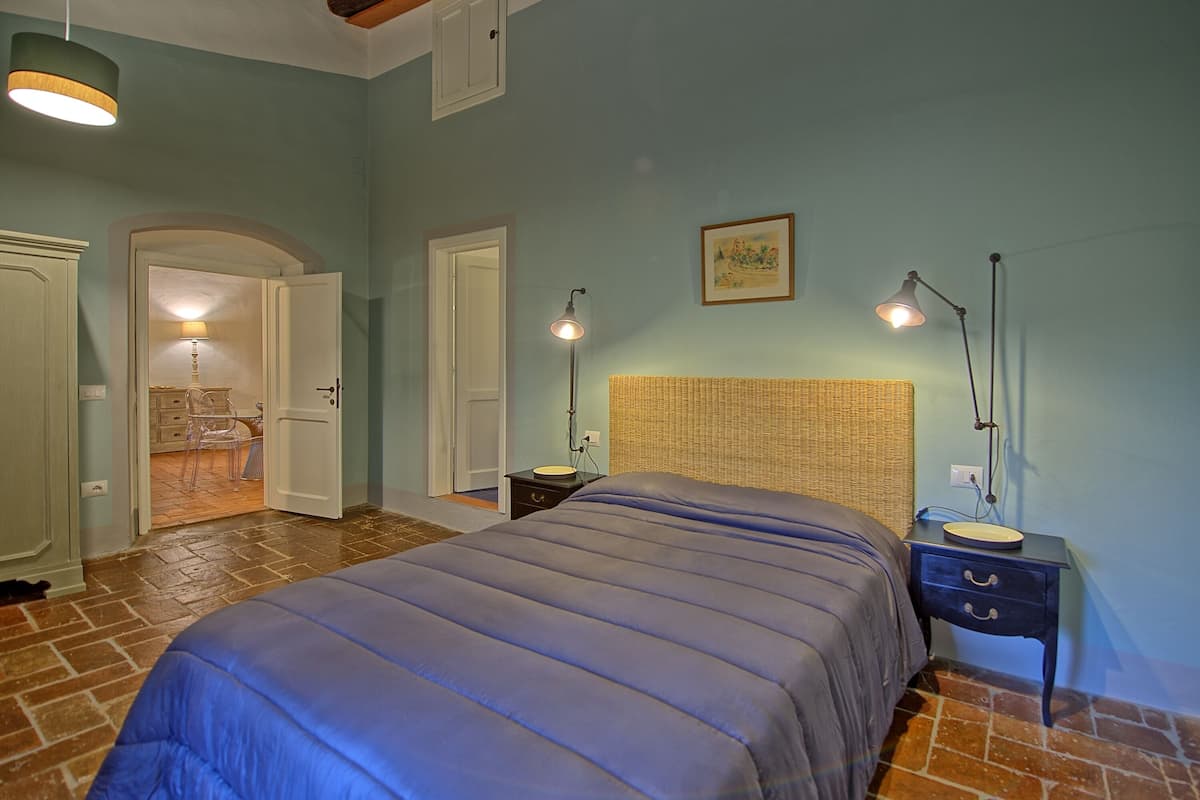 Quarto com uma cama grande, duas mesas de cabeceira, um criado-mudo, cabeceira de vime e uma porta que dá para um banheiro.