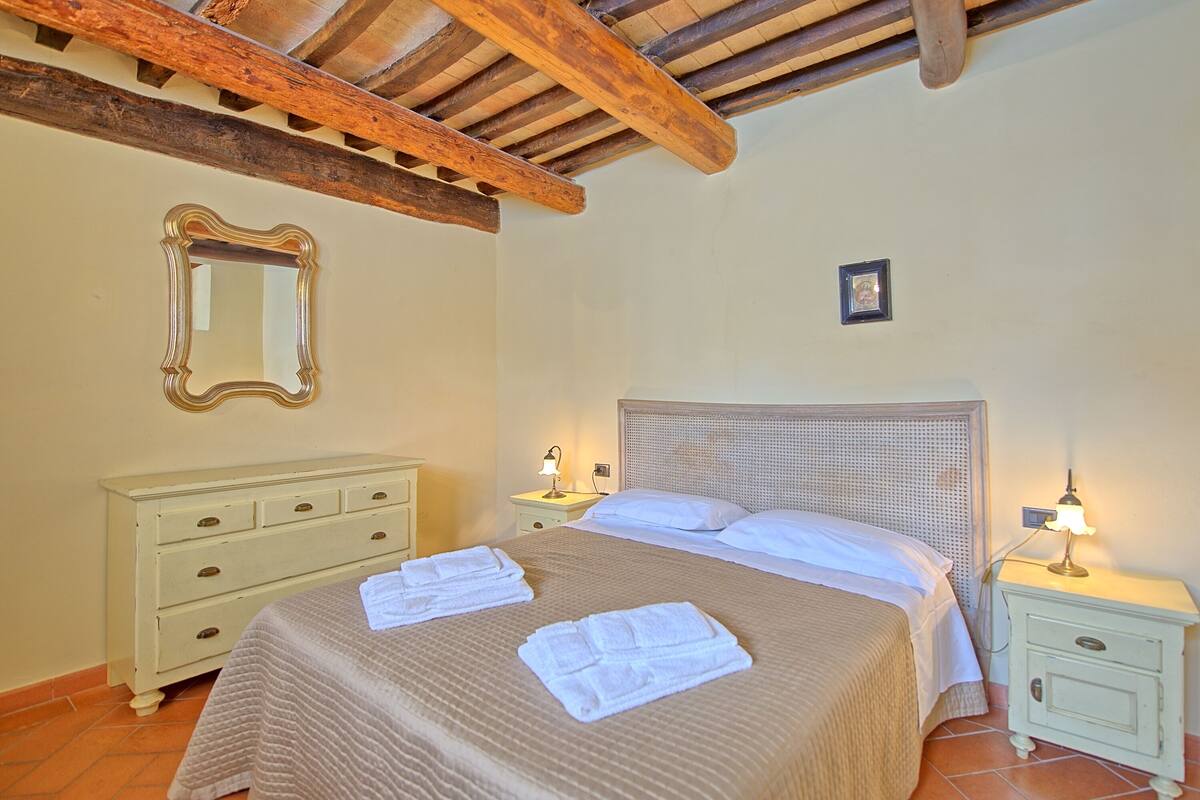 Quarto com cama, duas mesas de cabeceira, um criado-mudo, um espelho e teto com vigas aparentes.