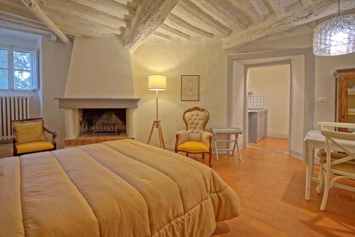 Quarto com cama grande, lareira, luminária de chão e poltrona com almofada amarela.
