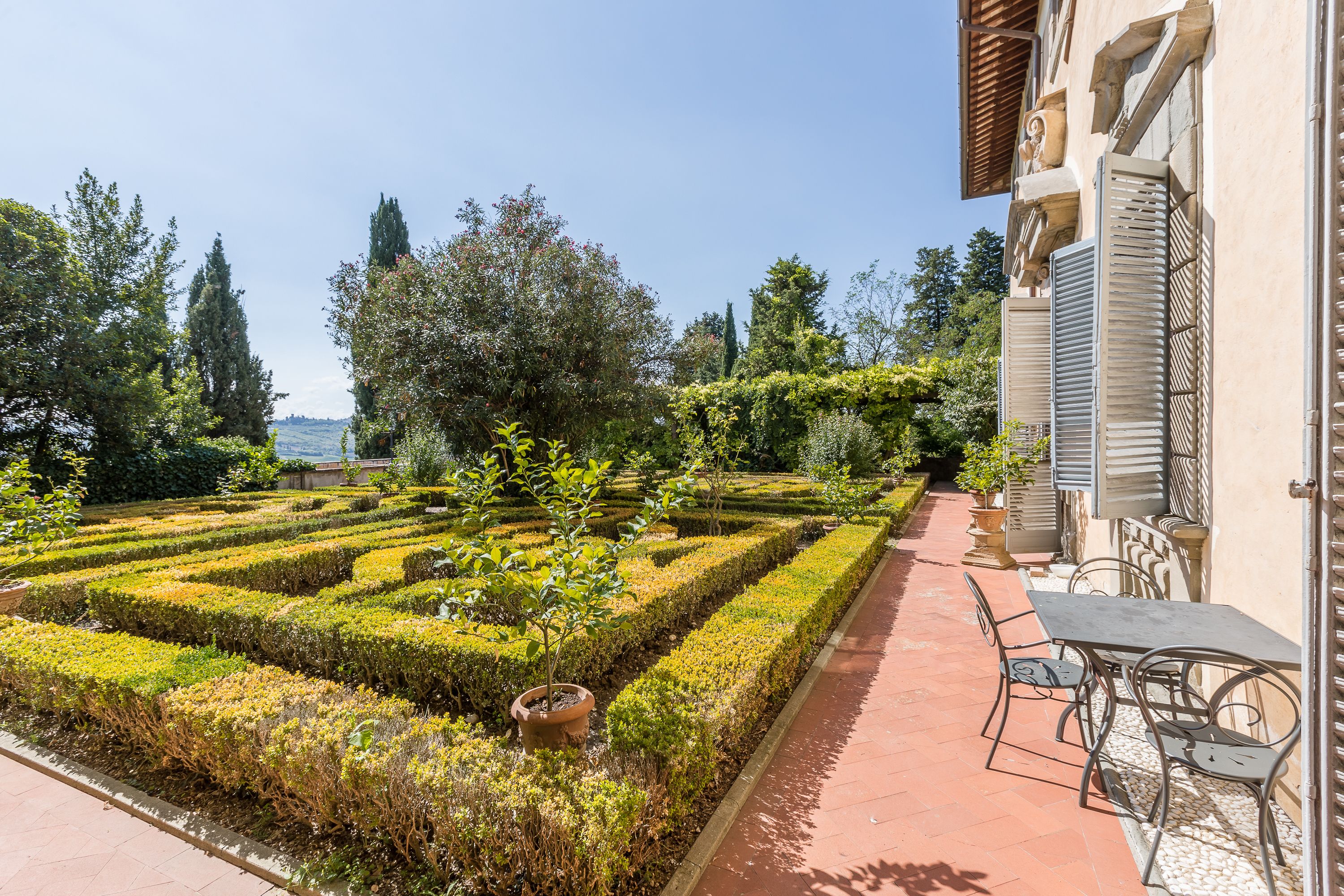 A garden with geometric hedges, a patio with a table and chairs, and a building with shutters.