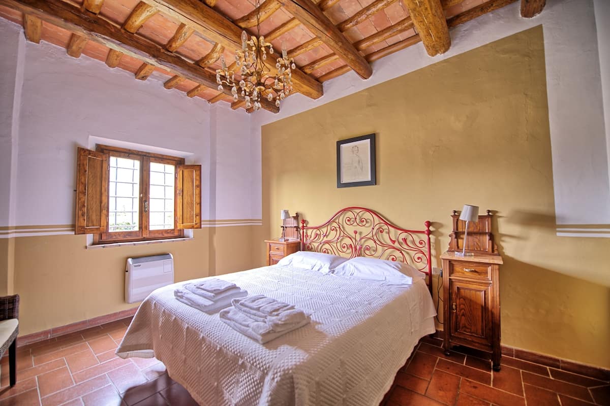 Quarto com cama, cabeceira de madeira, janela, radiador e mesa de cabeceira com abajur.