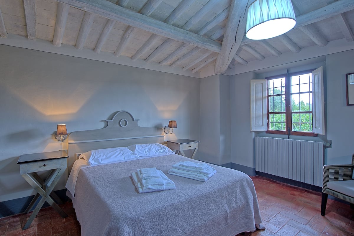 Quarto com uma cama, duas mesas de cabeceira com luminárias, uma janela com vista para o verde e um radiador.