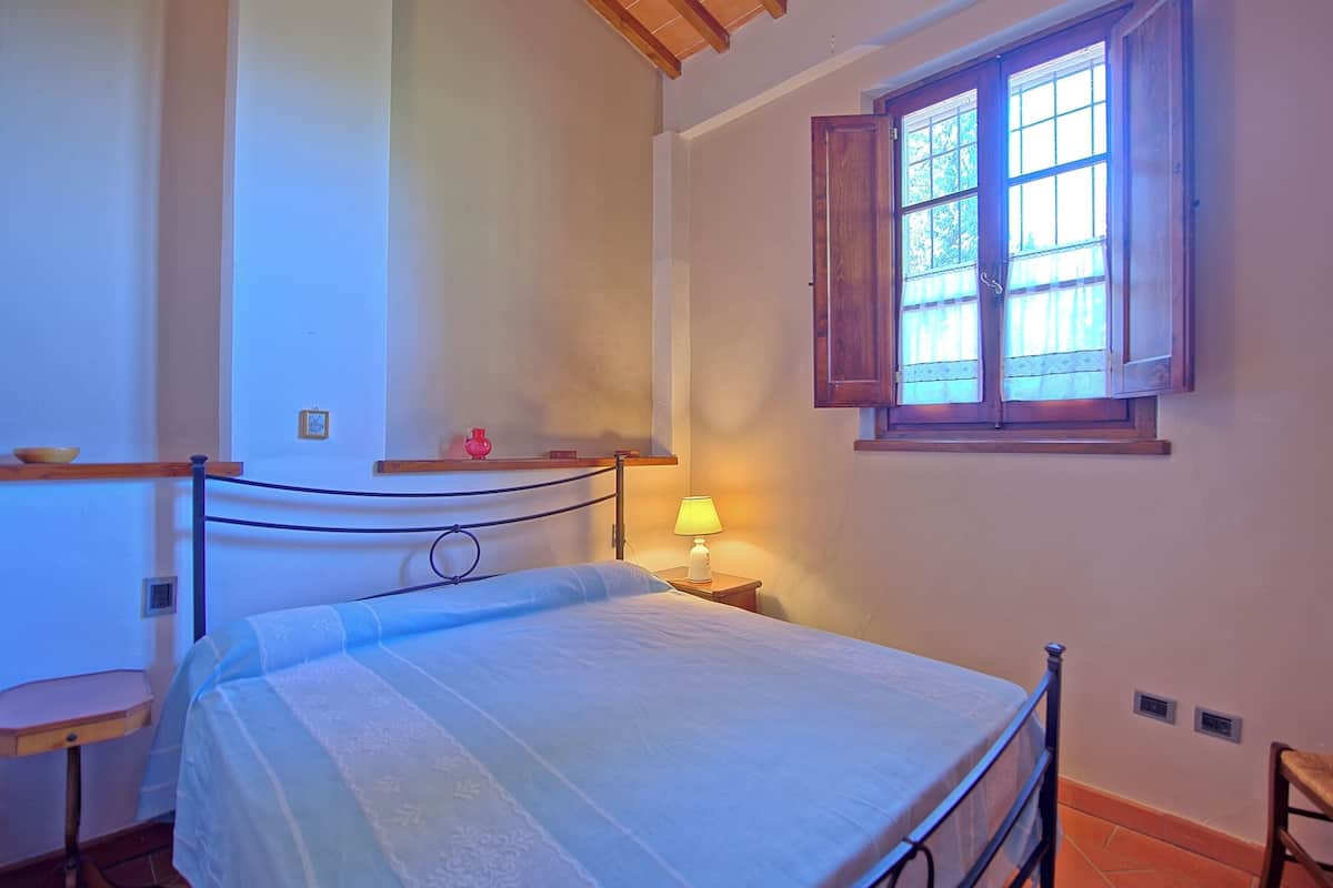 Um quarto com uma cama, uma mesa de cabeceira com abajur, uma janela com persianas de madeira e uma estante com uma tigela.