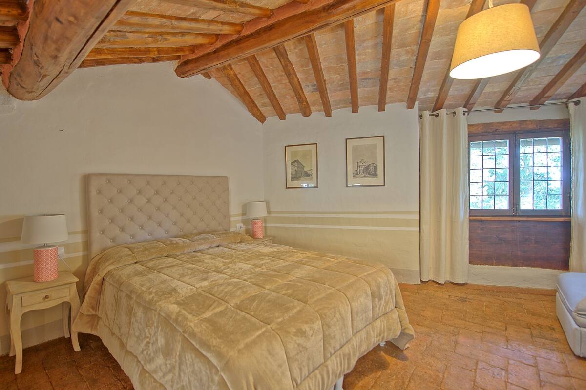 Um quarto com cama, duas mesinhas de cabeceira, luminária pendente e janela com cortinas.