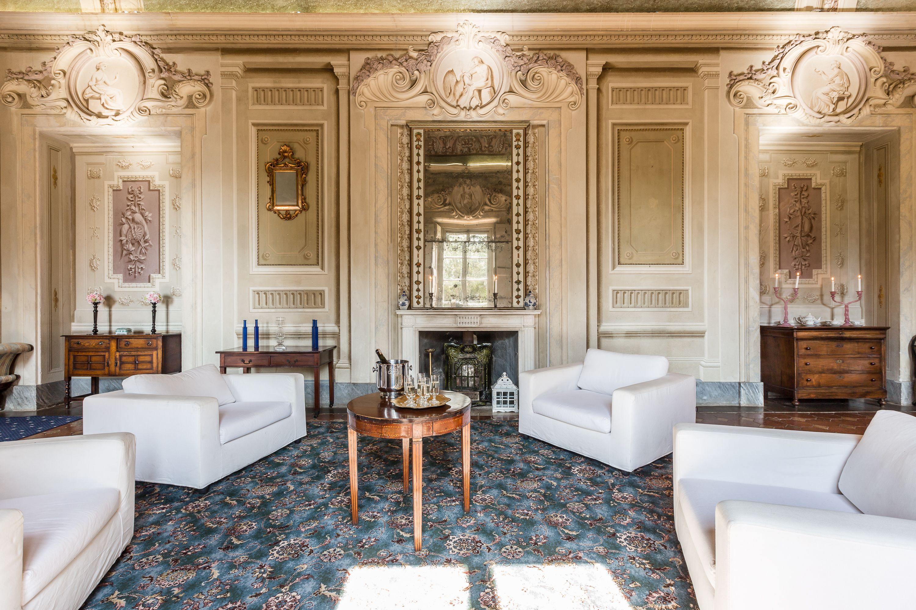 A grand room with ornate moldings, a fireplace, and white sofas arranged around a central table.
