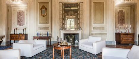 A grand room with ornate moldings, a fireplace, and white sofas arranged around a central table.