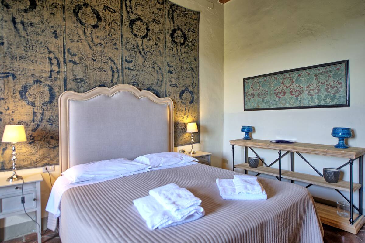 Uma cama bem arrumada com toalhas dobradas, uma mesa de cabeceira de madeira com uma luminária e uma parede com um quadro floral.