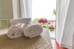 Une serviette blanche, soigneusement roulée, est posée sur un couvre-lit beige à texture marquée. De la terrasse, on aperçoit un immeuble et des plantes en pot.