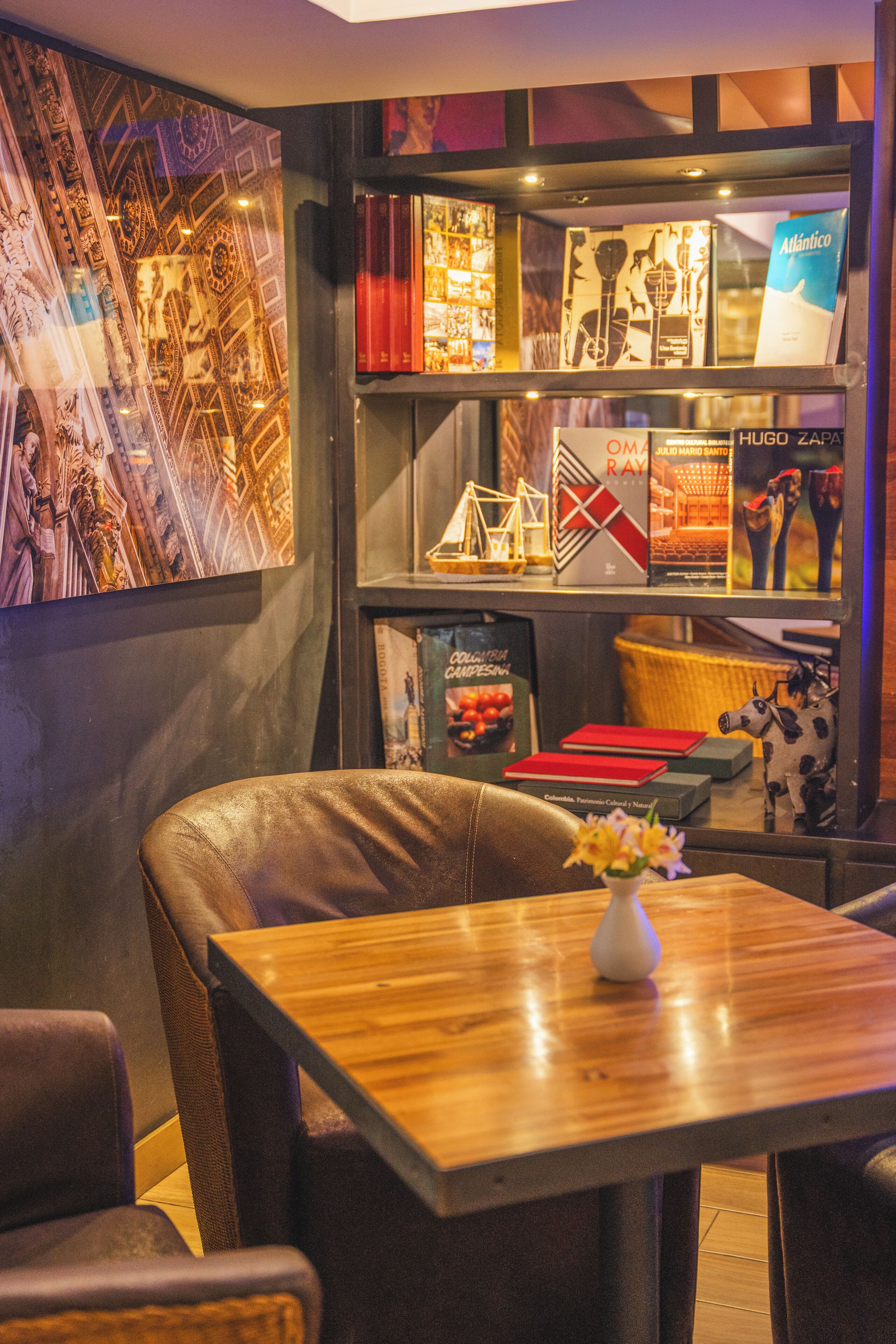 A cozy seating area with leather chairs and a wooden table, a bookshelf with books and decorative items, and a tapestry on the wall.