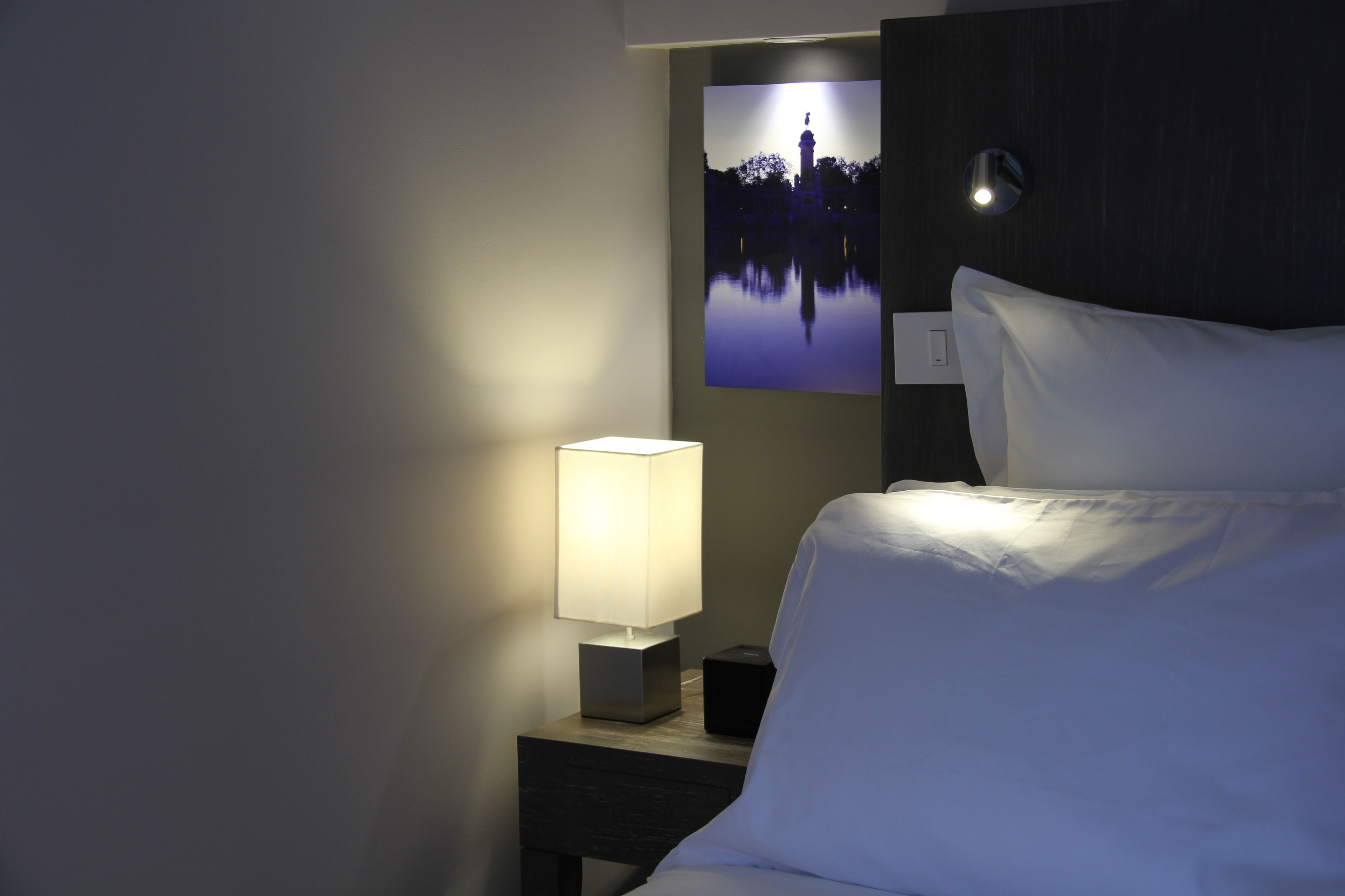 A hotel room with a bed, a bedside lamp, a nightstand, and a wall-mounted picture.
