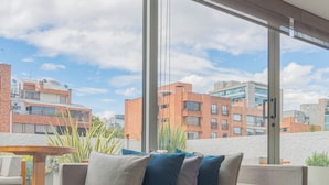 A modern living room with a beige sofa, a blue throw blanket, and a view of urban buildings through large windows. - GIO Hotel CITYFLATS Collection Bogotá (Bogotá)