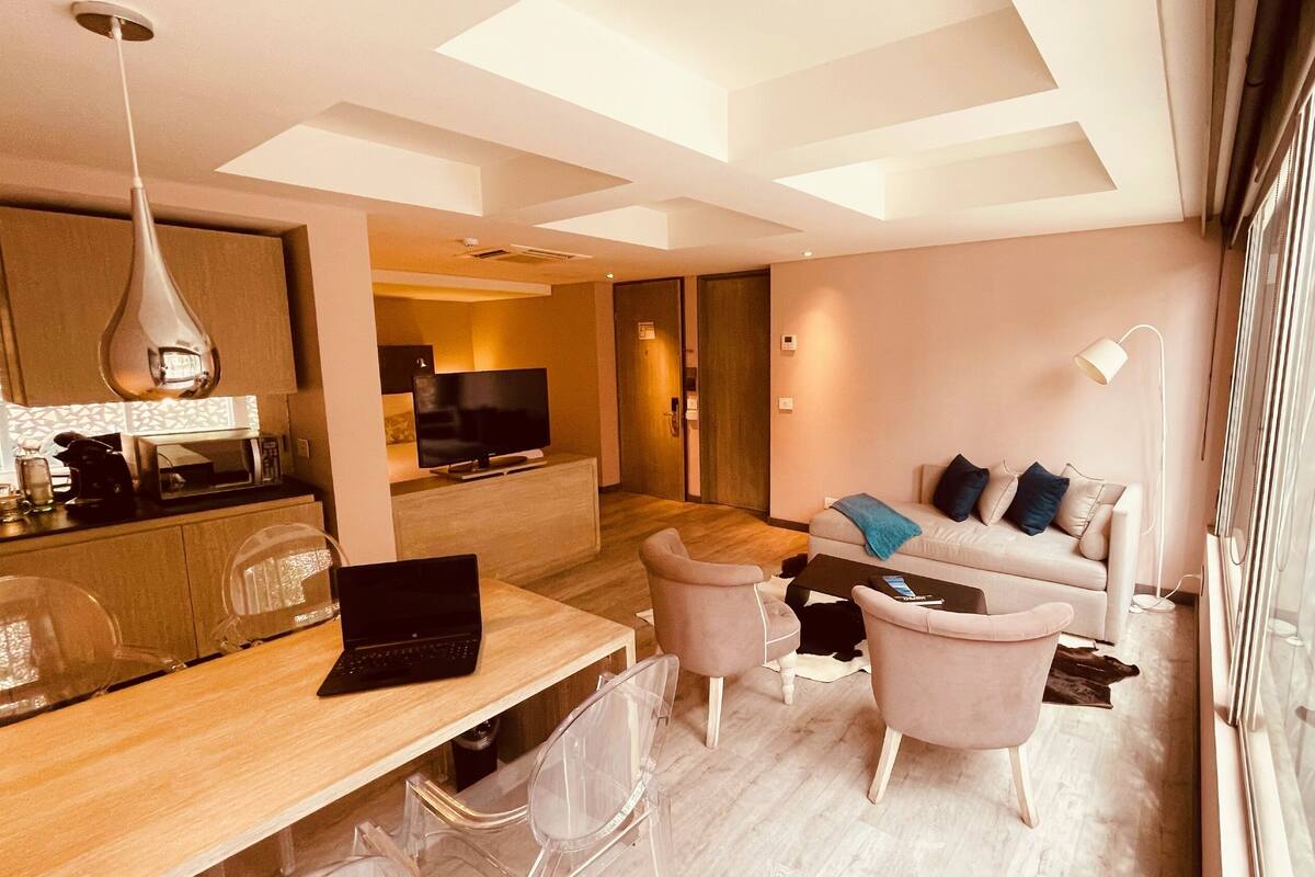 A modern living room with a sofa, chairs, a coffee table, a TV, and a dining area with a laptop and a coffee machine.