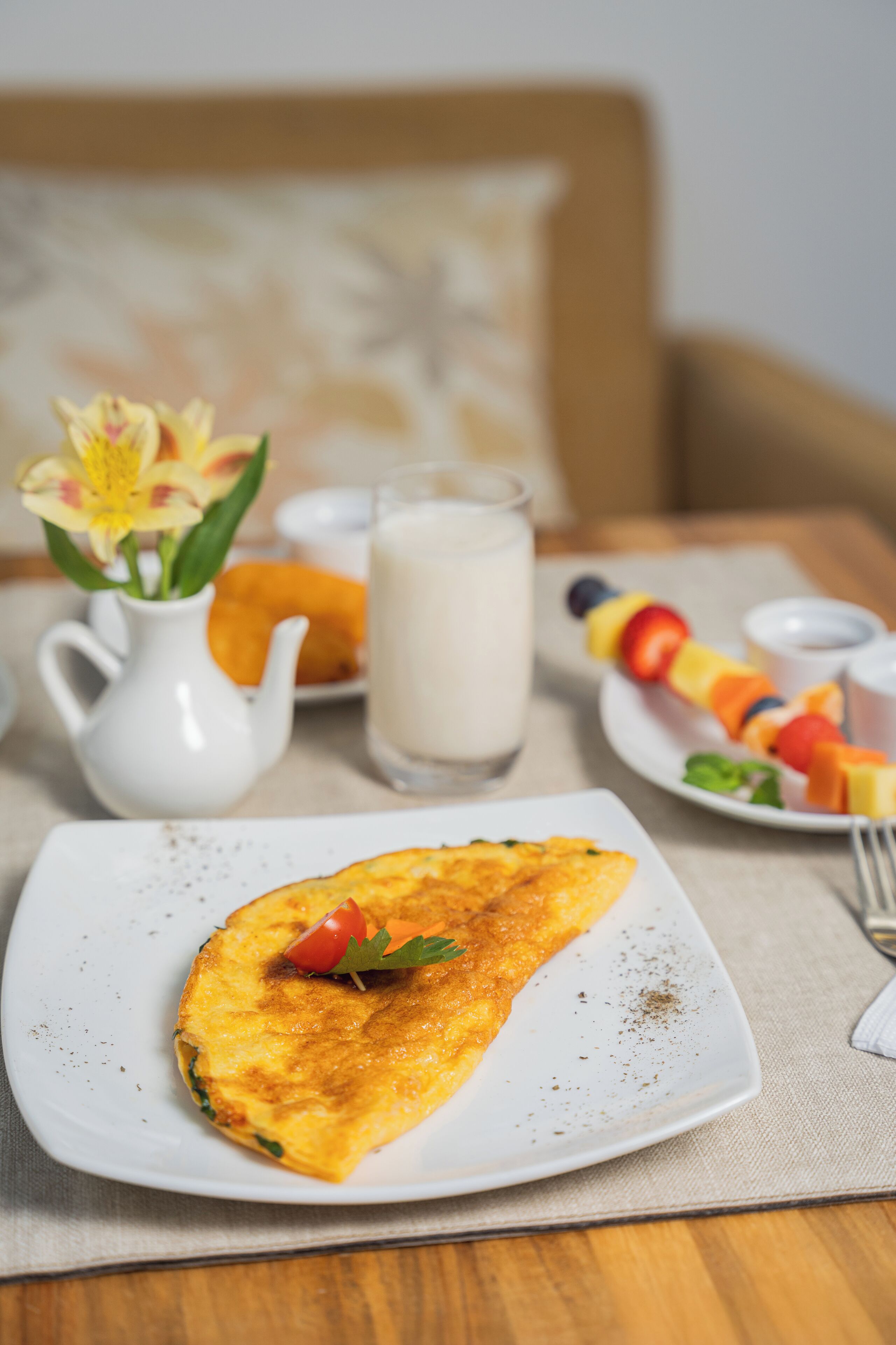 A breakfast setting with an omelette on a plate, a pitcher of milk, and a fruit skewer on a table.