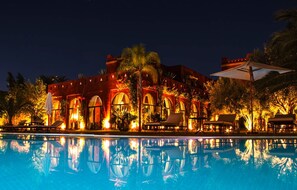 Ein Hotel bei Nacht mit Schwimmbecken, das Gebäude und die umliegenden Bäume spiegelt.