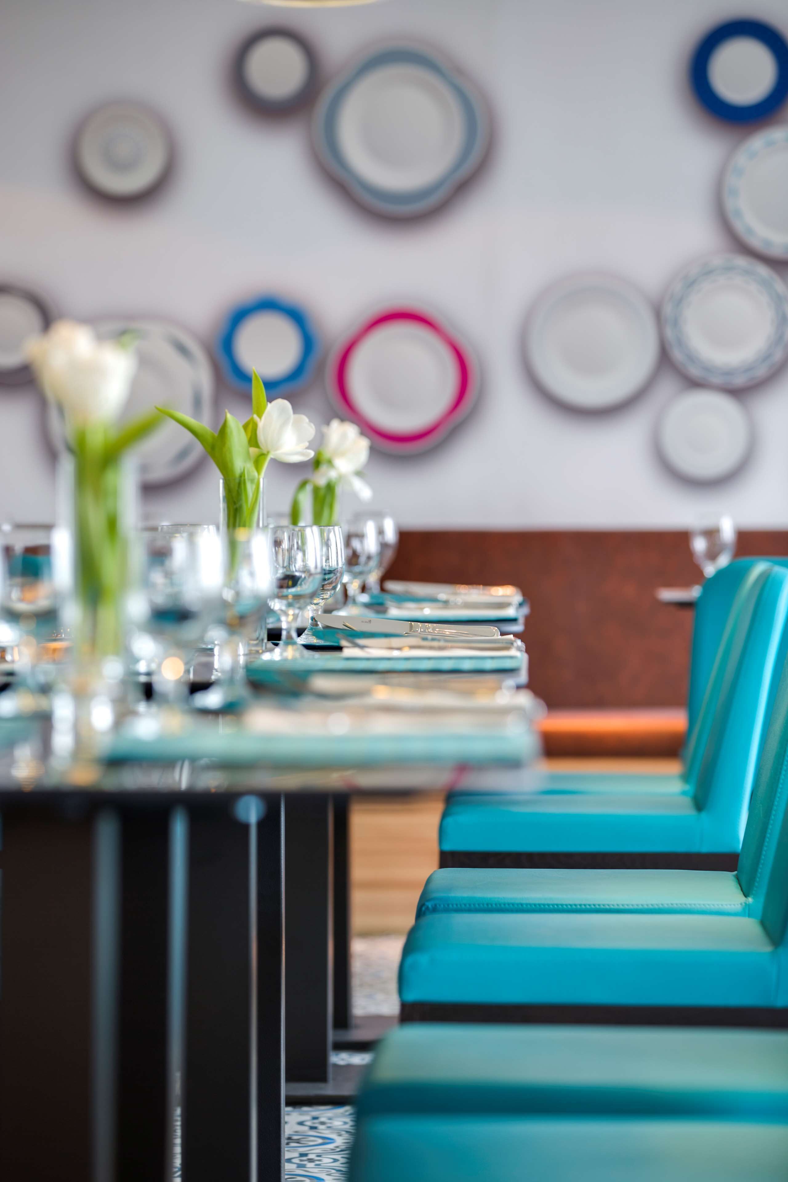 A dining area with teal upholstered chairs, a long table set with plates, glasses, and a vase with white flowers.