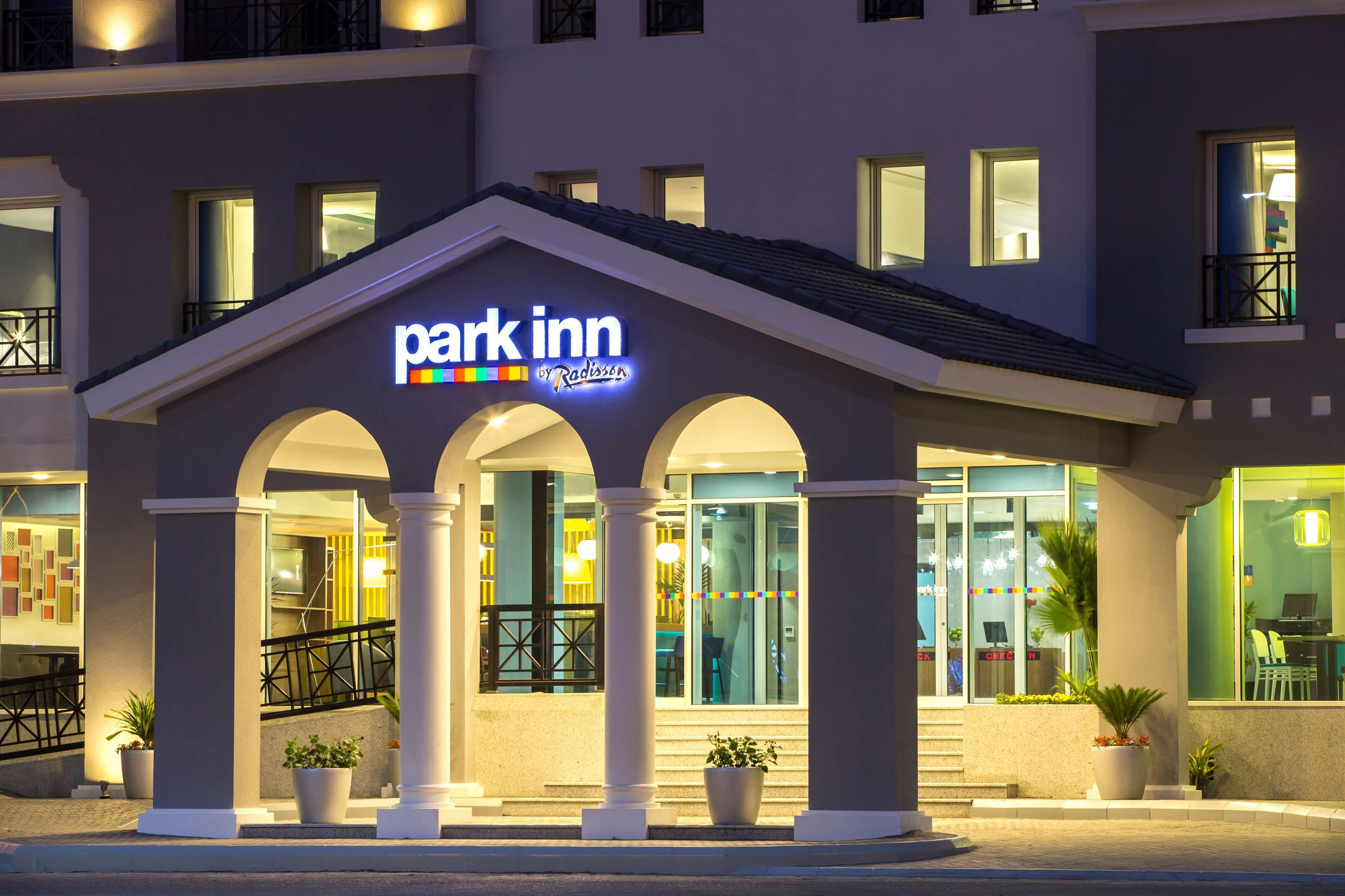 A hotel entrance with a 'Park Inn by Radisson' sign, featuring a modern facade with large windows and a central archway supported by columns.