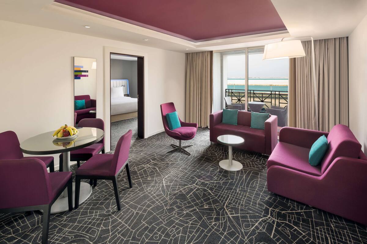 A modern hotel room with a bed, purple and teal furniture, a dining table with fruit, and a view of the beach.