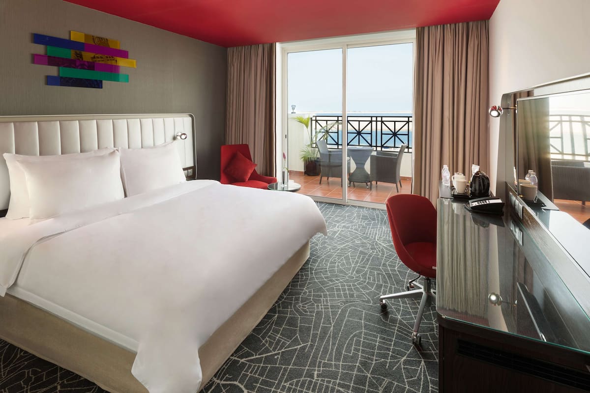 A hotel room with a large bed, a desk with a TV, a red chair, and a balcony with a view of the sea.