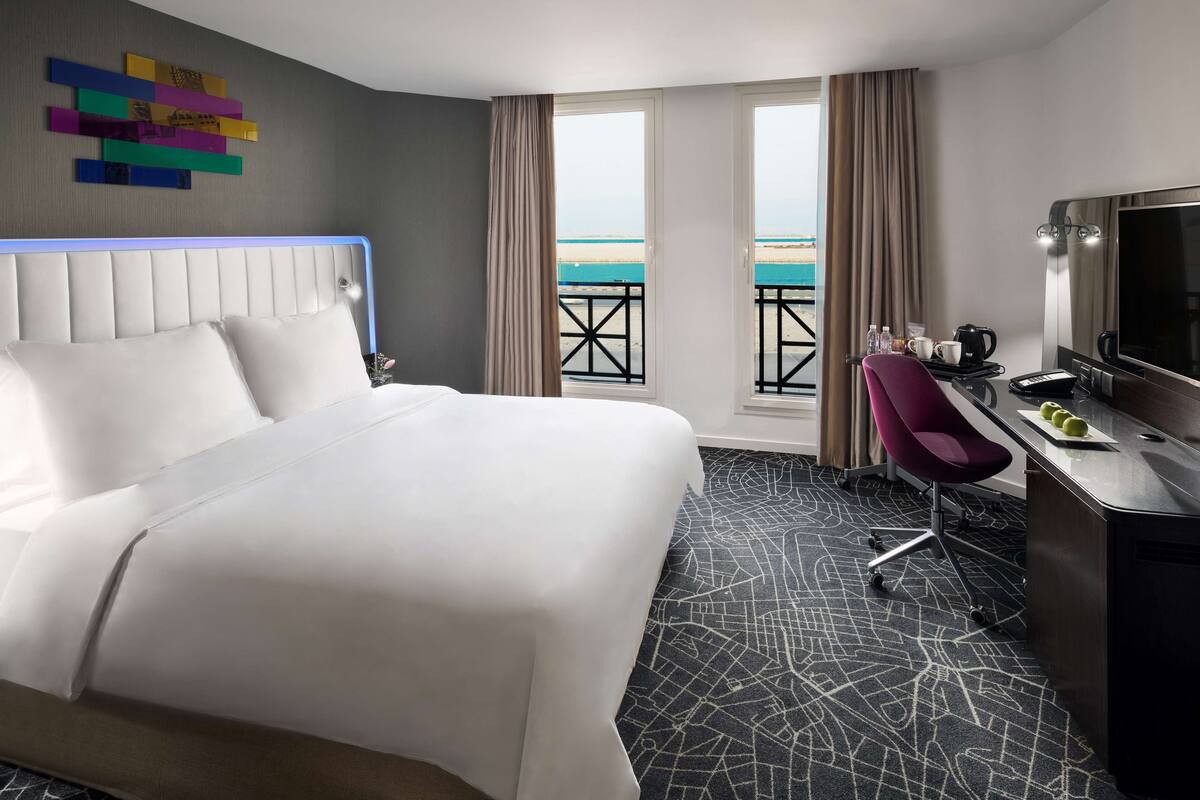 A hotel room with a large bed, a desk with a chair, a television, and a view of the beach through the window.