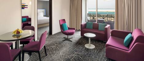 Habitación de hotel con cama, sillones morados, una pequeña mesa con fruta, un balcón con vista a la playa y una alfombra estampada.