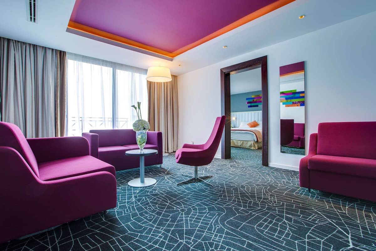 A modern hotel room with a purple color scheme, featuring a sofa, armchair, and a small table with a vase.