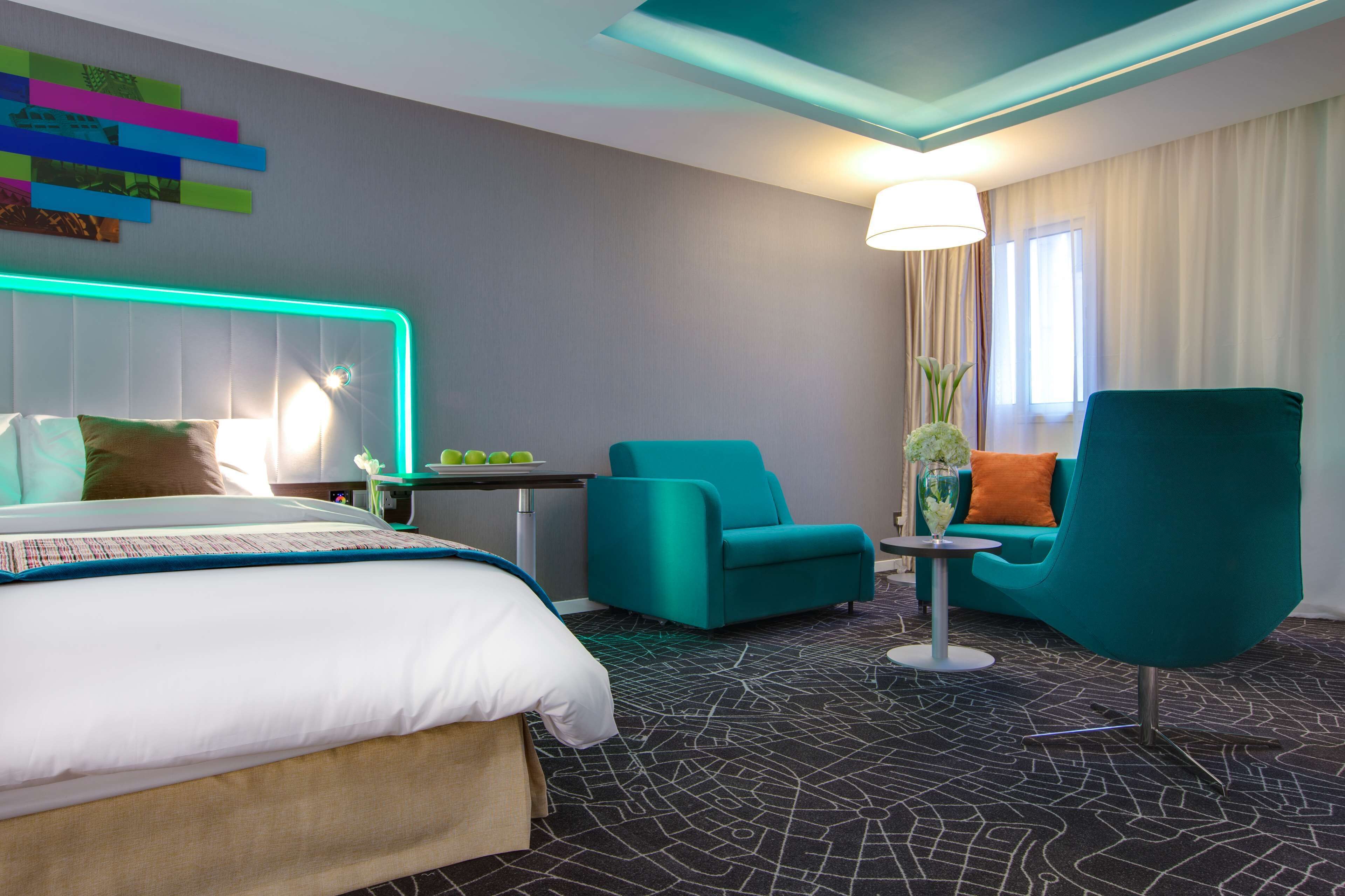 A hotel room with a bed, two teal armchairs, a small table, a lamp, and a decorative wall piece.