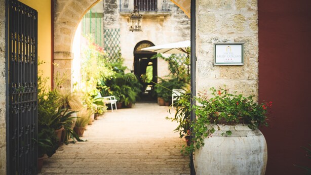 An arched entrance to a courtyard with potted plants, a sign for Hotel Baguio Catalano, and a view into the building. - Hotel Baglio Catalano (Custonaci)