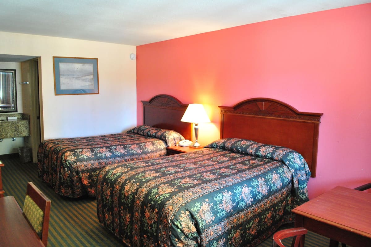 Une chambre d’hôtel avec deux lits, une tête de lit en bois, une table de chevet avec une lampe, un tableau encadré au mur et un bureau avec une chaise.