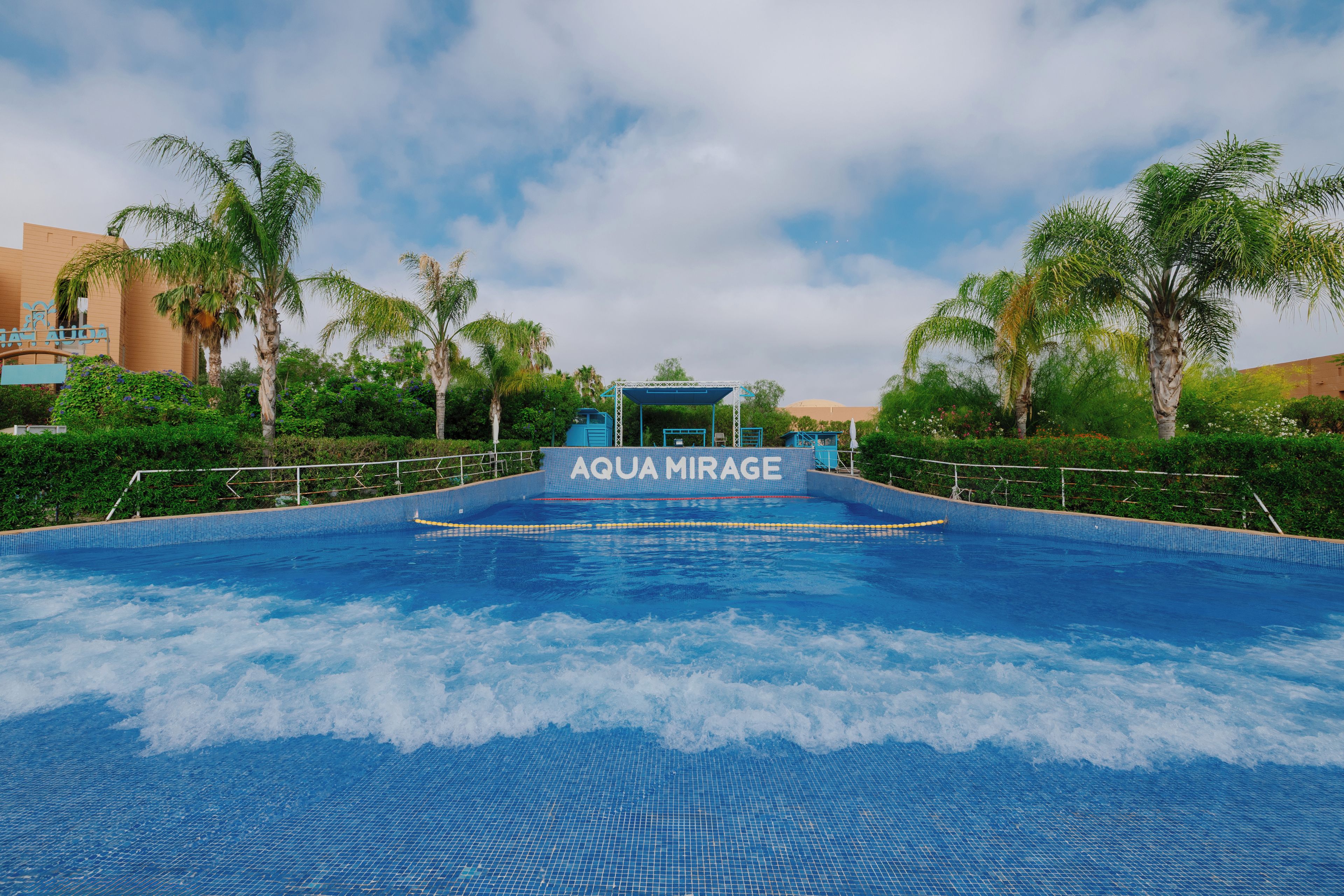 An indoor swimming pool with a sign reading 'AQUA MIRAGE' and a view of the surrounding greenery and buildings.