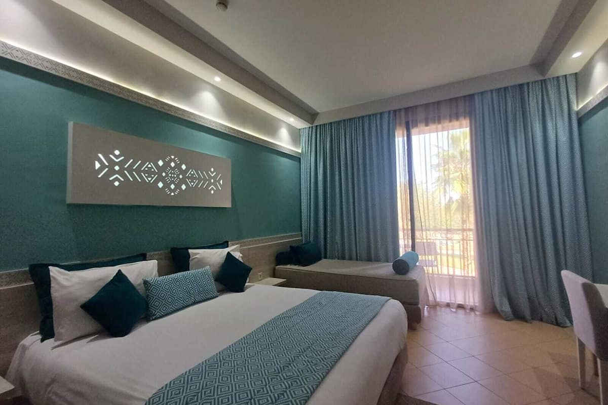 A hotel room with two beds, a large window with curtains, a wall-mounted artwork, and a ceiling with recessed lighting.