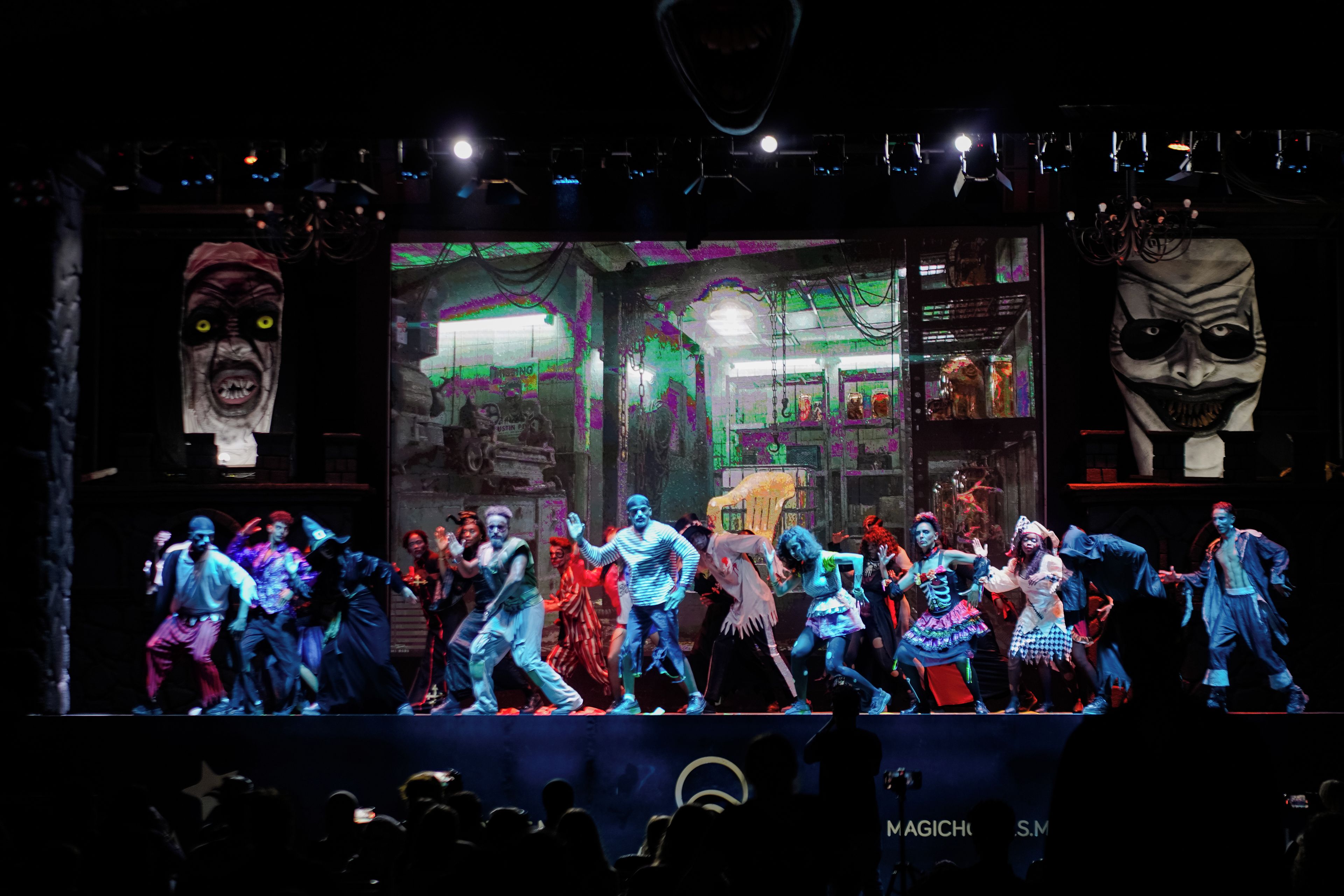A stage with performers in costumes, a backdrop with graffiti, and a crowd in the foreground.