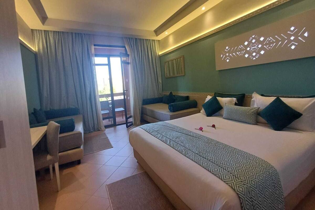 A hotel room with a large bed, two armchairs, a small table, and a view through a sliding door to an outdoor area.