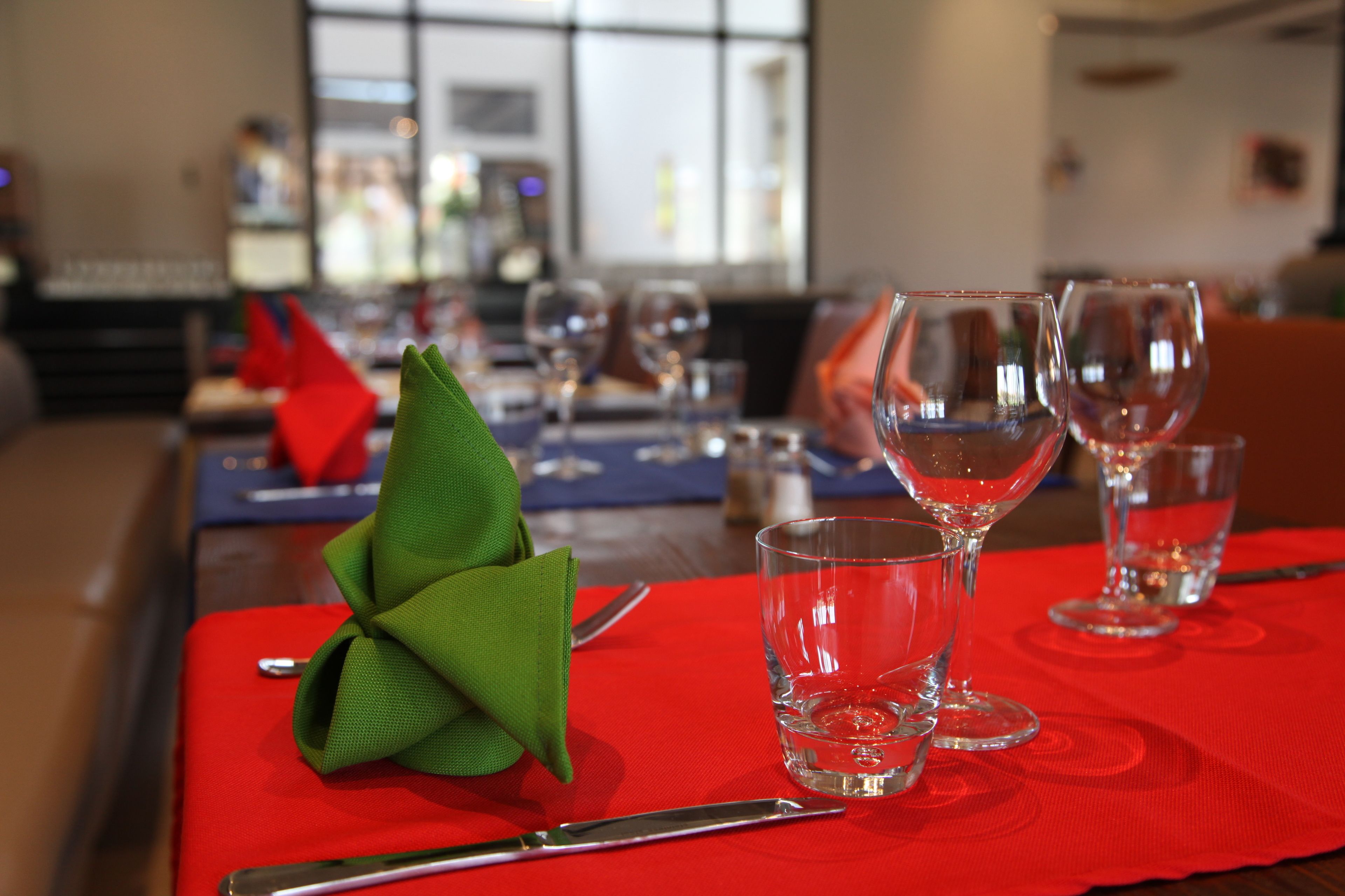 Une table à manger dressée avec des sets de table rouges, des verres à vin transparents et une serviette verte.