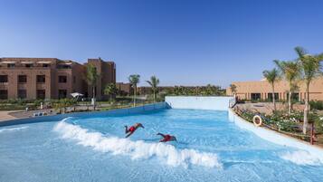 Una piscina con dos personas en trajes de baño rojos, rodeada de palmeras y un edificio de techo plano.